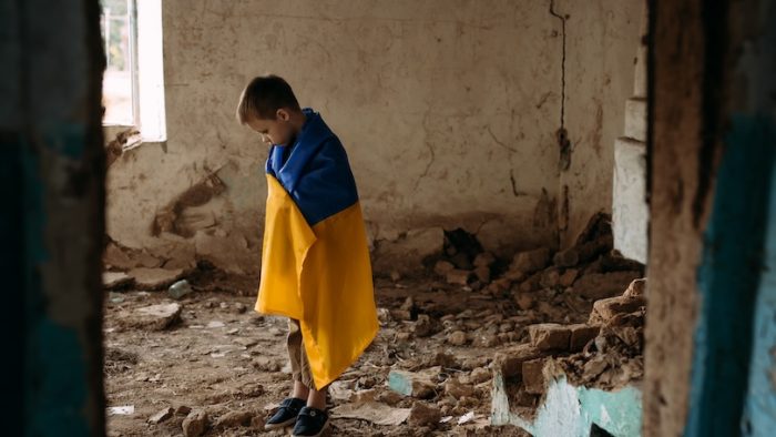 A child stands in a dilapidated house and looks around. A boy wrapped in a Ukrainian flag stands among the ruins. Blurry background and copy space for your advertising text message.