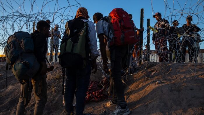 Migrants with backpacks await entry at the barbed-wire Ciudad Juarez border during sunset.
